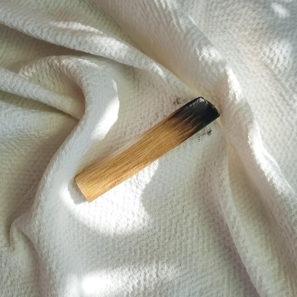 Un batonnet de bois de santal est déposé sur une serviette ondulée blanche. C'est la fragrance Soleil Noir de Erra. Disponible en diffuseur et bougie.