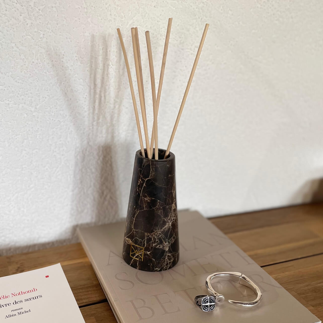 Diffuseur de parfum en marbre noir avec bâtonnets de rotin, déposé sur une petite table de chevet, à côté d'un livre et de bijoux en argent.