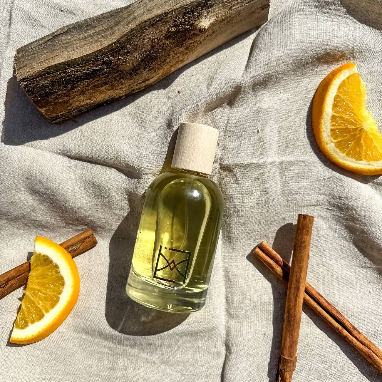 Bouteille de parfum d'ambiance écologique avec bouchon en bois, entourée de tranches d'orange, de bâtons de cannelle et d'un morceau de bois flotté sur un tissu en lin.