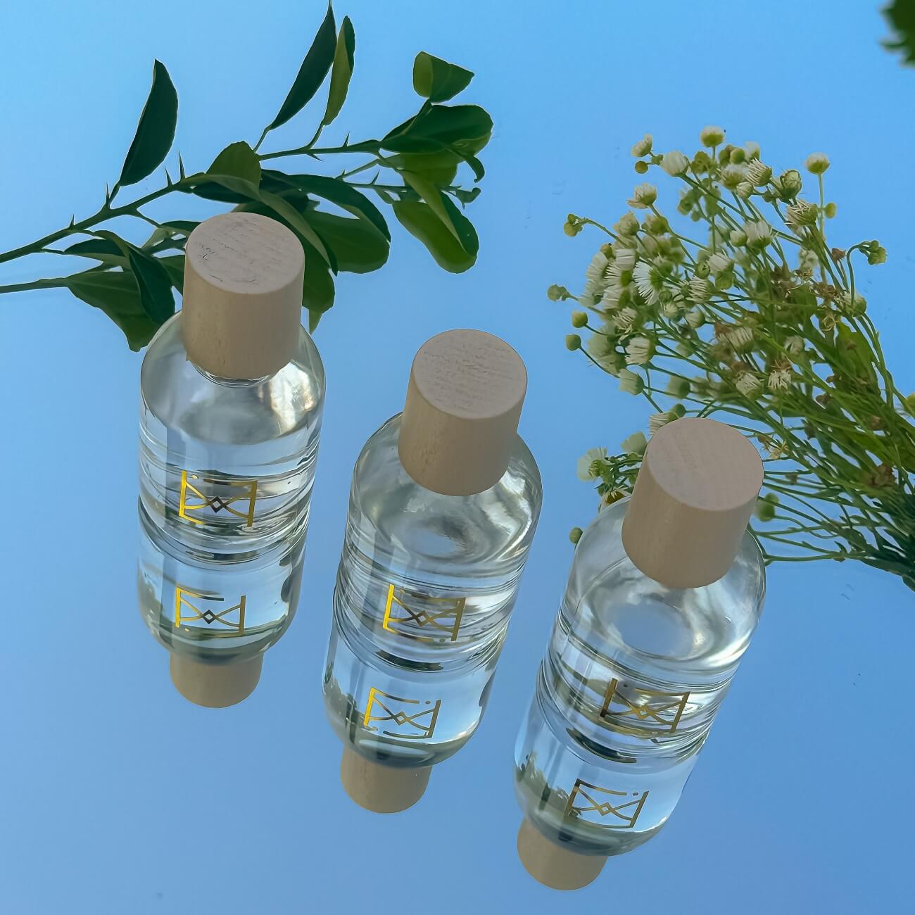 Trois bouteilles de parfum d'ambiance écologique avec bouchons en bois, entourées de verdure et de fleurs blanches sur fond bleu.